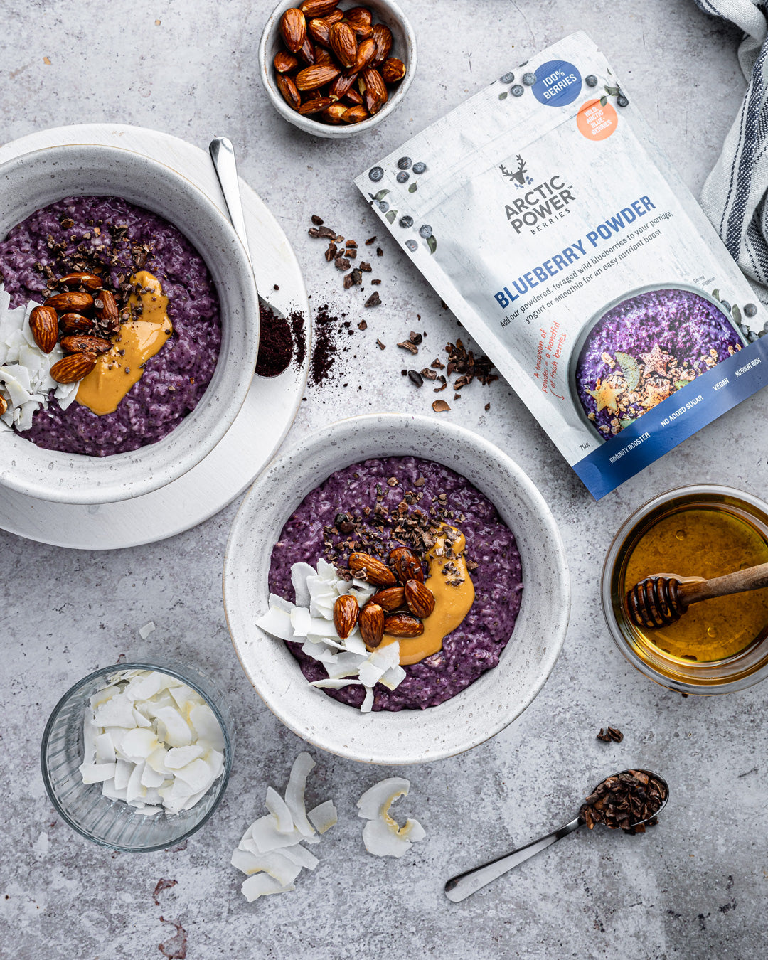 Two bowls of dark blue porridge with toppings on a gray surface, featuring Arctic Power Berries Blueberry powder packaging.