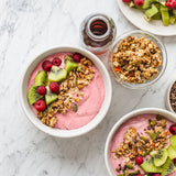 Two bowls of raspberry powder yoghurt with granola, kiwi, and raspberries on a marble surface.
